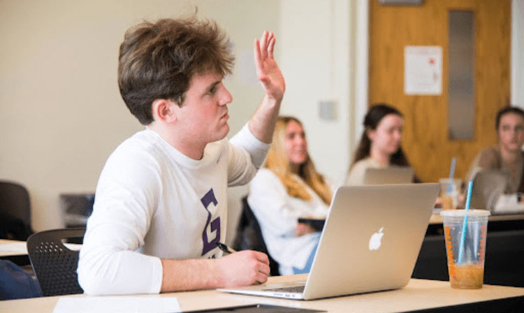 A student raises his hand in front of an open laptop.