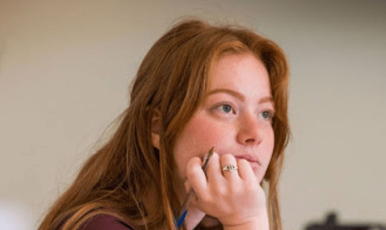 A student holds a pen in her hand, resting her chin on her fist, and looks forward.