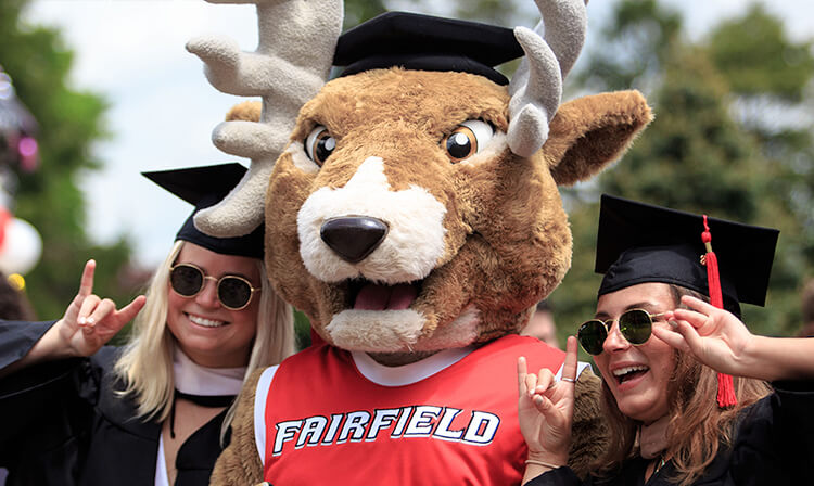 Two women in graduation gowns and caps pose joyfully with a Lucas the Fairfield University mascot, celebrating their academic achievement together.