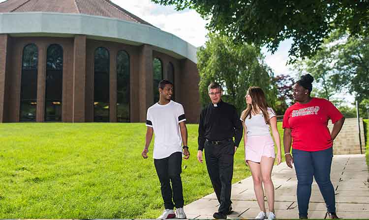 3 students walk past chapel with priest