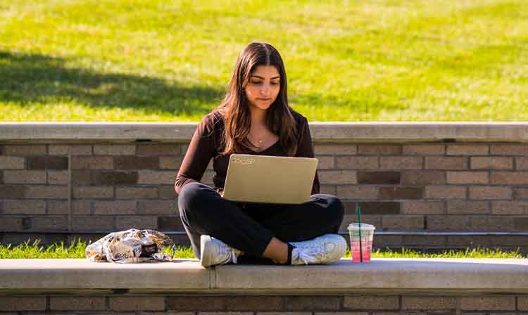 A student sits on a sitting wall outdoors with their laptop on their lap.