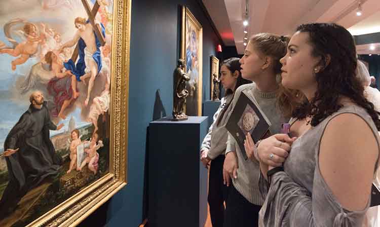 Art History students admiring a painting portraying Ignatius  staring up at Jesus as he ascends with a cross surrounded by angels. 