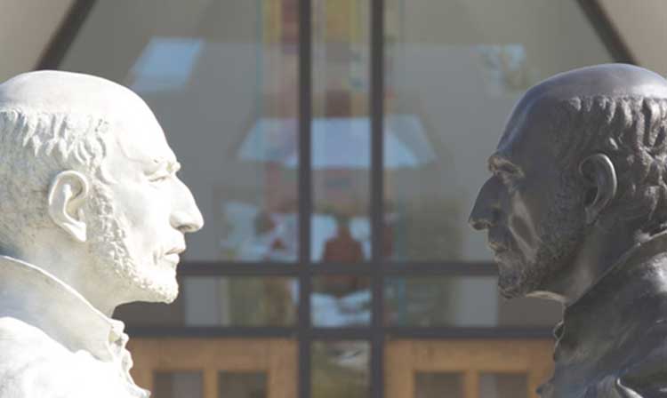 Two sculptures of Ignatius on campus in front of the chapel facing each other with an intense gaze. One sculpture is white and one is significantly darker. 