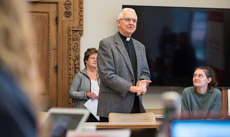 A priest standing teaching a class with a professor standing next to a screen behind him on his right and a student looking at him sitting on his left. 