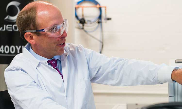 A Chemistry professor wearing a lab coat and protection pointing to a screen emphasizing something on it. 