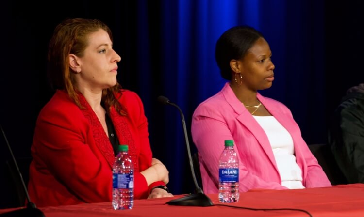 Two professors sitting on a panel behind a red table with water bottles on top of it. 