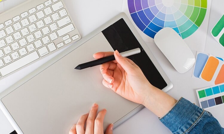 A person holds a digital pencil over a drawing pad with paint sachets surrounding a keyboard. 