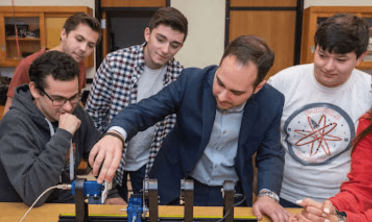 A professor touches a machine while a group of students surround and watch.