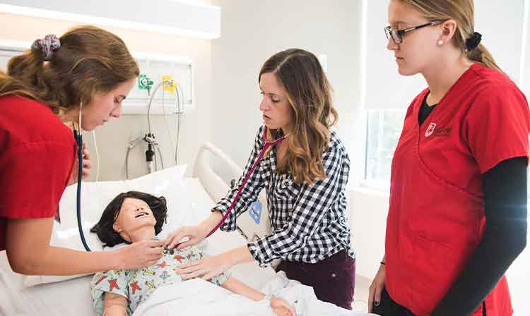 Two students in red scrubs watch as a professor uses a stethoscope on a manikin.