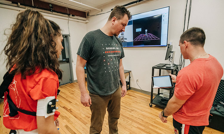 Professor observes students screen as they stand in project room. 