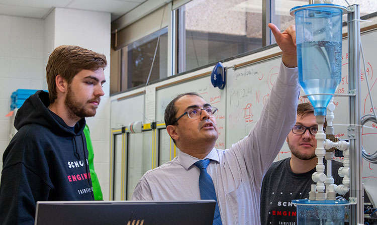 Professor reaches up to something on a machine with two students observing. 