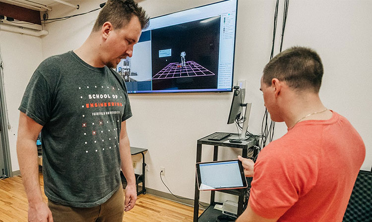 Two men stand together, looking at a computer screen, engaged in discussion or analysis.