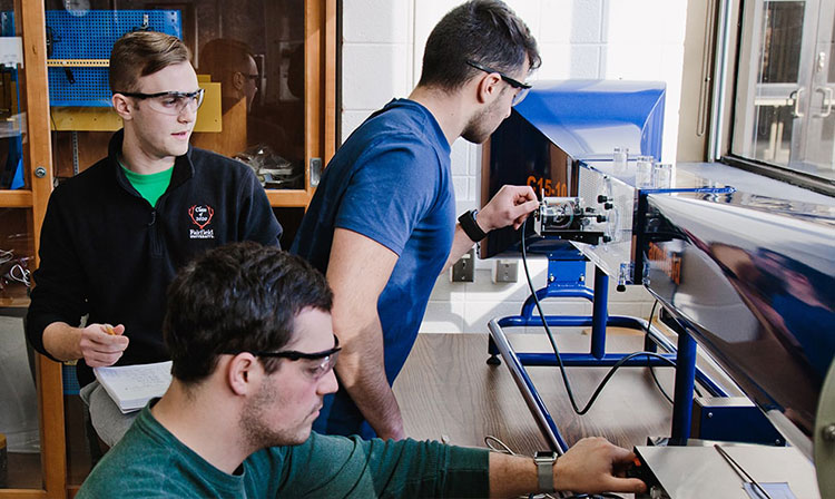 Three men in a lab collaborating on a machine, focused on their work and surrounded by tools and equipment.