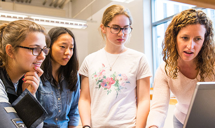 Four women gather around a laptop, focused and engaged in discussion as they explore content on the screen.