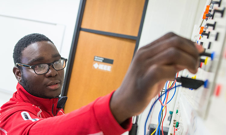 A young man wearing a red shirt is intently working on a circuit board, surrounded by tools and components.