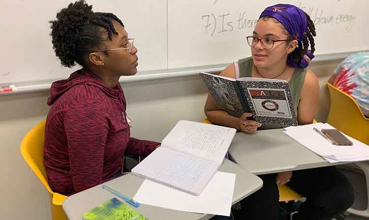 2 students sit near white board and discuss classwork 