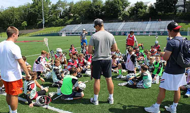 3 Lacrosse coaches talk to group of campers sitting on field 