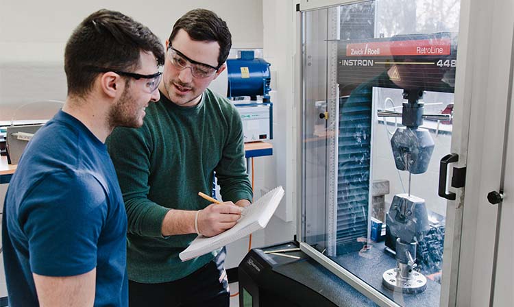 Students wearing eye protection looking over a machine that says “Instron” on it. 