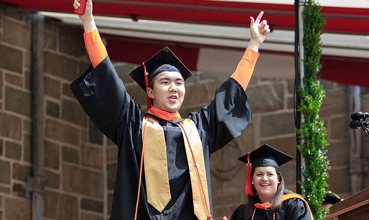 A man in a graduation gown and cap joyfully raises his arms in celebration of his achievement.