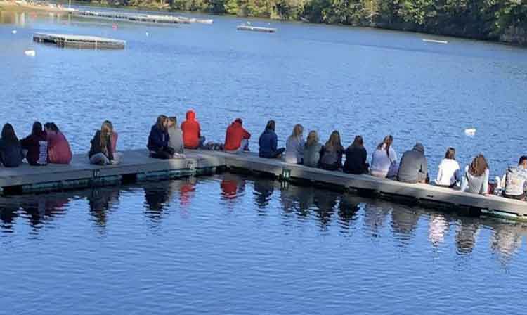 A group of students sit on a dock outdoors overlooking a blue lake.