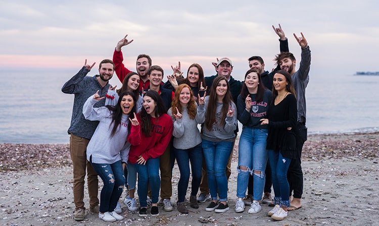 A group of students stand on a beach in front of a pink and purple sunset.