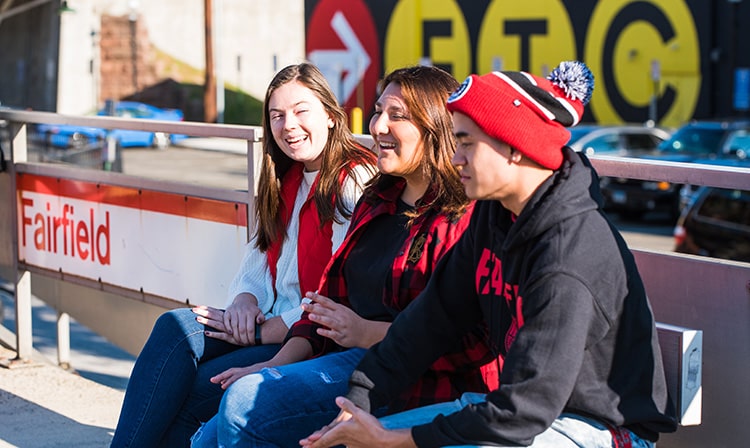 Three students sit at a bench on a train platform with a sign that reads “Fairfield”.