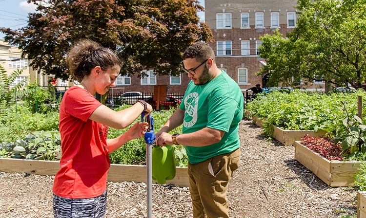 Two people fill a plastic container with water from a spout outdoors.