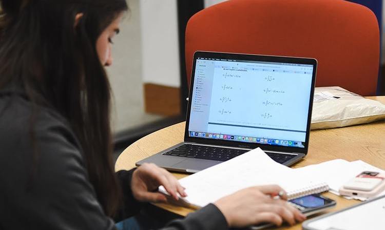 A student uses a calculator and notebook in front of an open laptop displaying math equations