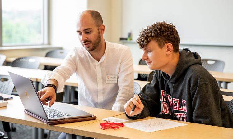 A student and professor sit at a desk looking at an open laptop.