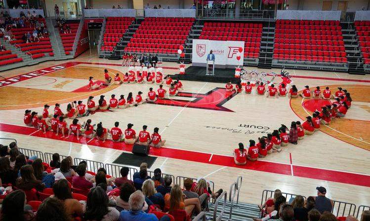 Students sit on the floor of a gymnasium in the shape of a 27.