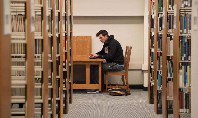 A student works at a study carrel in a library, seen through an aisle of shelves