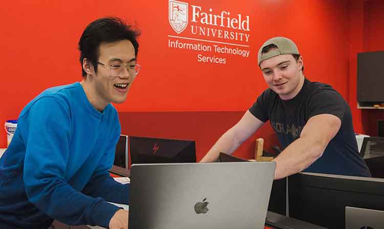 Two people discuss an open laptop in front of a red wall that reads “Fairfield University Information Technology Services”. 