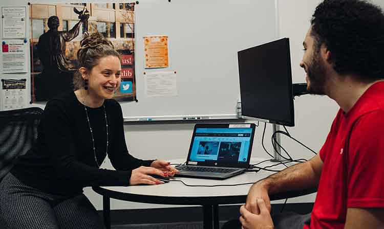 Two people talk in front of an open laptop and a monitor not in use.