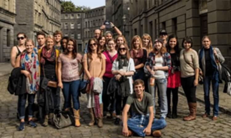 A large group of students standing in the middle of a cobblestone street in a foreign place. 