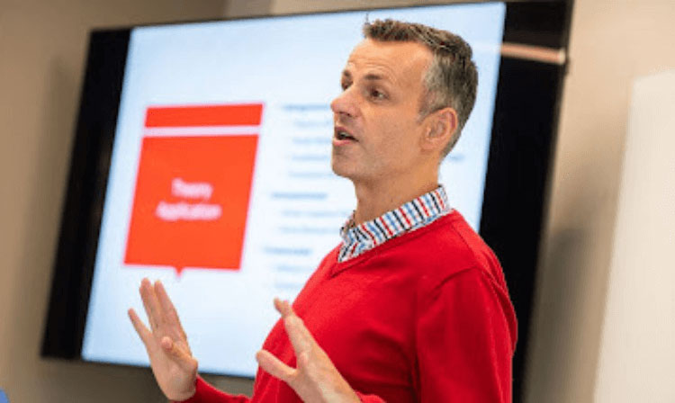 A professor in a red sweater stands in front of a PowerPoint addressing a class.