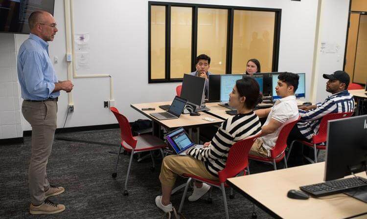 A professor leads a class of graduate students seated at a table with laptops, engaging in an engineering lectur.