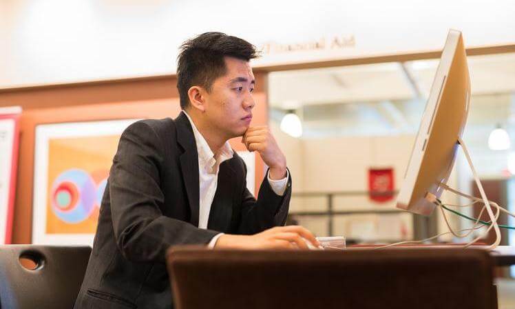 A graduate student is focused on reading on a large computer screen. He is dressed in a gray sport coat and white shirt.