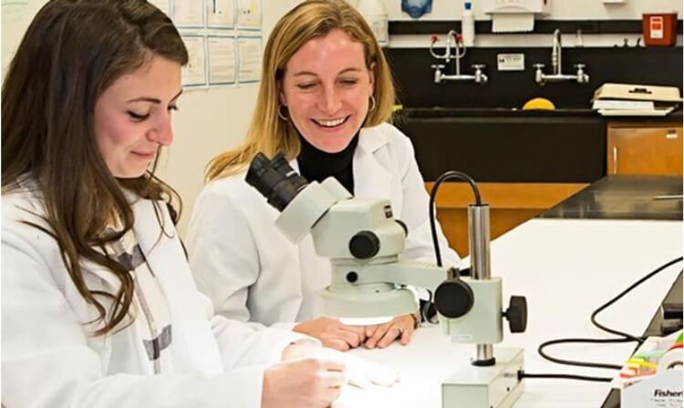 Two students sit at a desk in lab coats with a microscope in front of them.