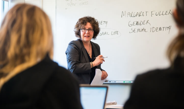 A professor stands in front of a whiteboard holding a piece of paper and an expo marker, facing the class.