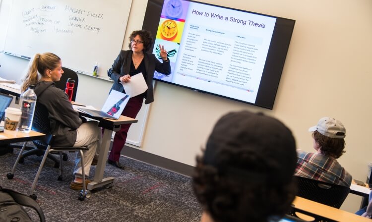 Professor teaches students in a small classroom emphasizing a screen behind her that reads “How to write a strong thesis”. 