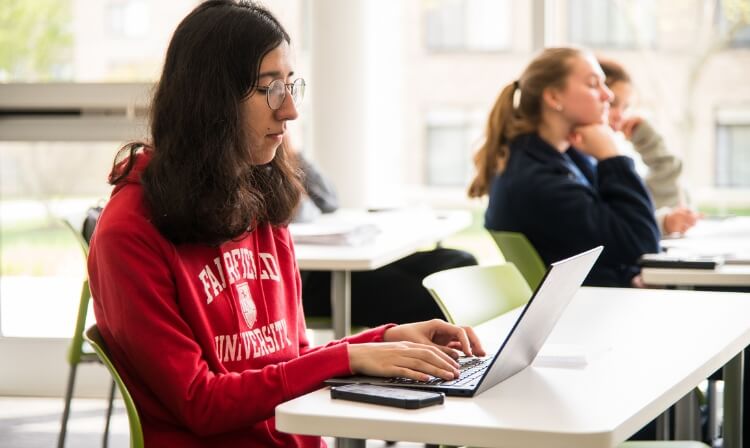 A student in a Fairfield University sweatshirt sits at a desk on their laptop.