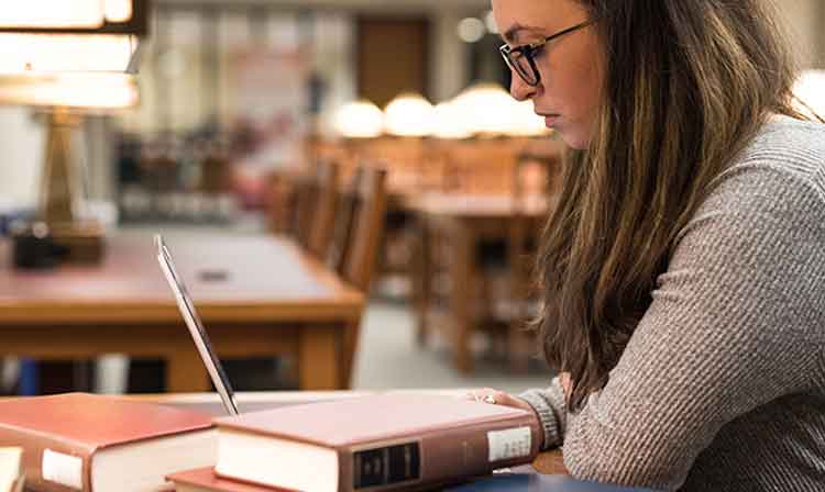 A student works on laptop in the library next to a stack of books.