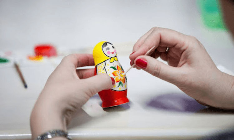 A person uses a toothpick to decorate a Russian doll with yellow paint.