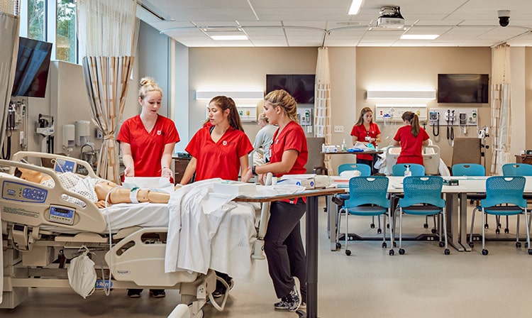 Students in red scrubs work with two different mannequin in a simulation room.