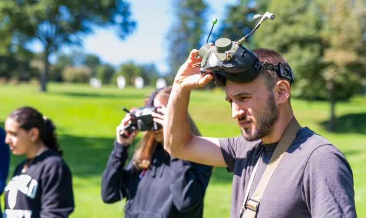 Engineering professor wearing FPV goggles. He is standing outside with two other students on bright green grass. 