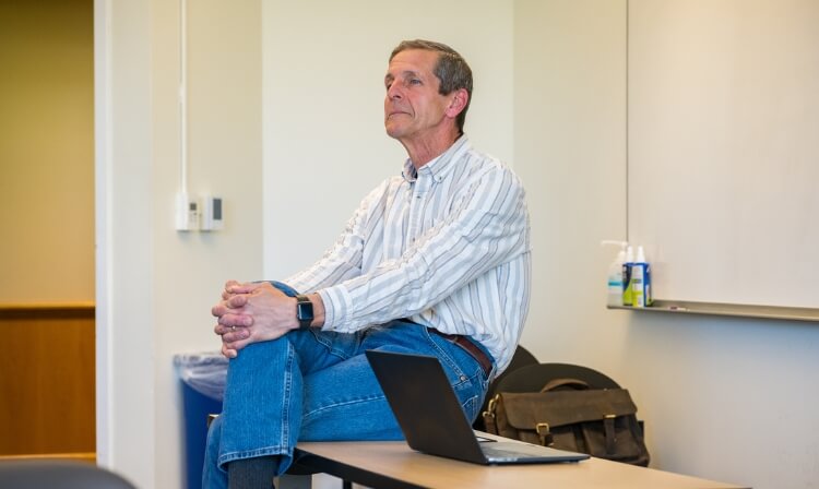 A faculty member sits on a desk in a classroom.