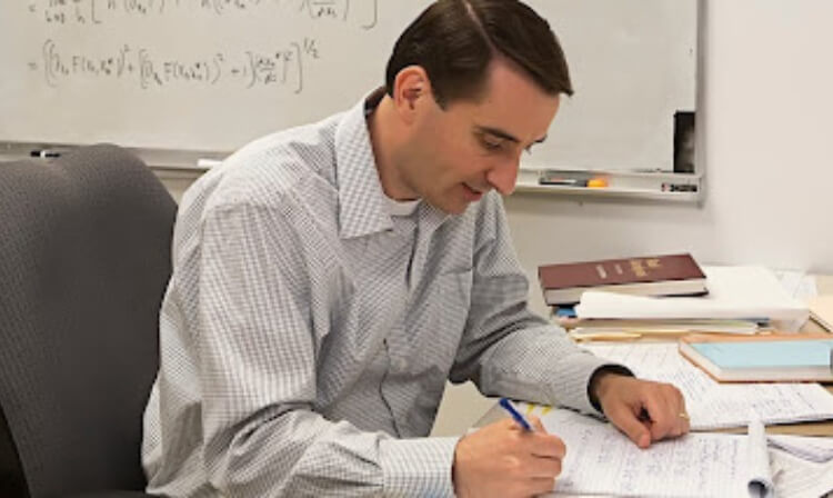 A professor sits at a desk looking at equations on a sheet of paper.