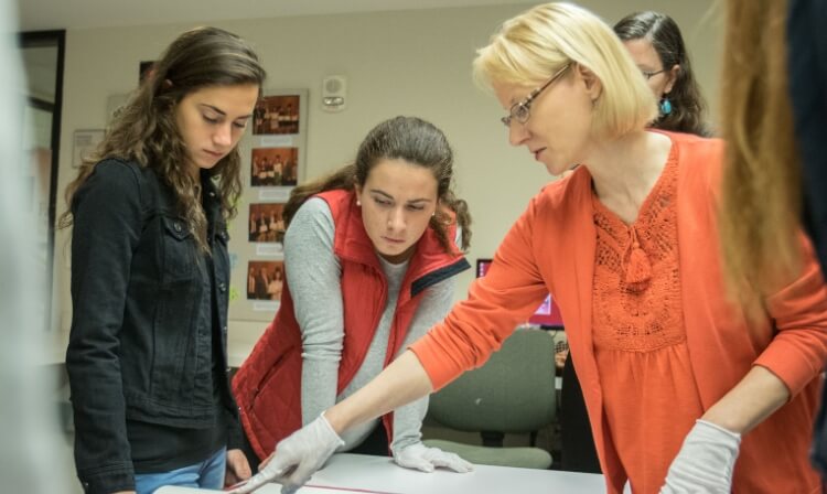 A professor wearing gloves points at a painting laying on a table in front of two students.