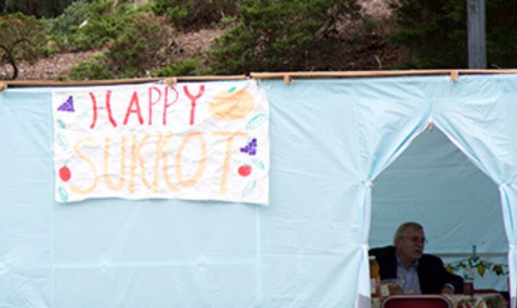 A blue tent outdoors has a sign that reads “Happy Sukkot”. 