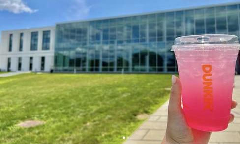 A person holds up a pink drink from Dunkin’ in front of a glass building.